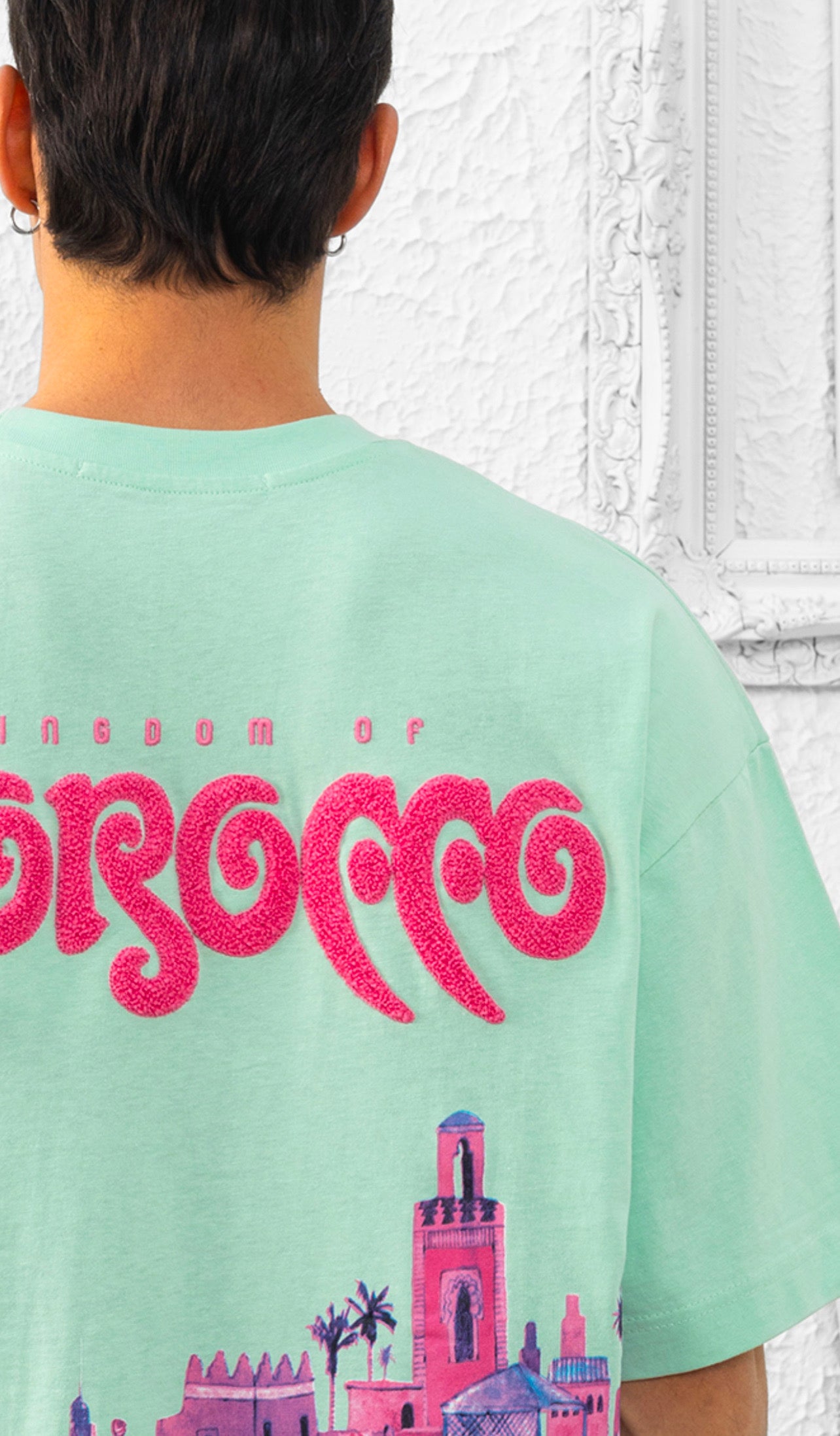 T-shirt oversize couleur mint avec illustration rose inspirée du Maroc et lettrage brodé Morocco, porté par un homme devant un mur texturé blanc.
